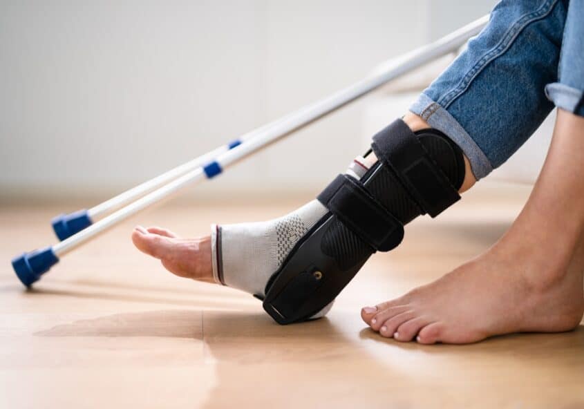 Persona que lleva una tobillera negra y una bota ortopédica en el pie lesionado, y que utiliza muletas como apoyo mientras se recupera de un esguince o una fractura.