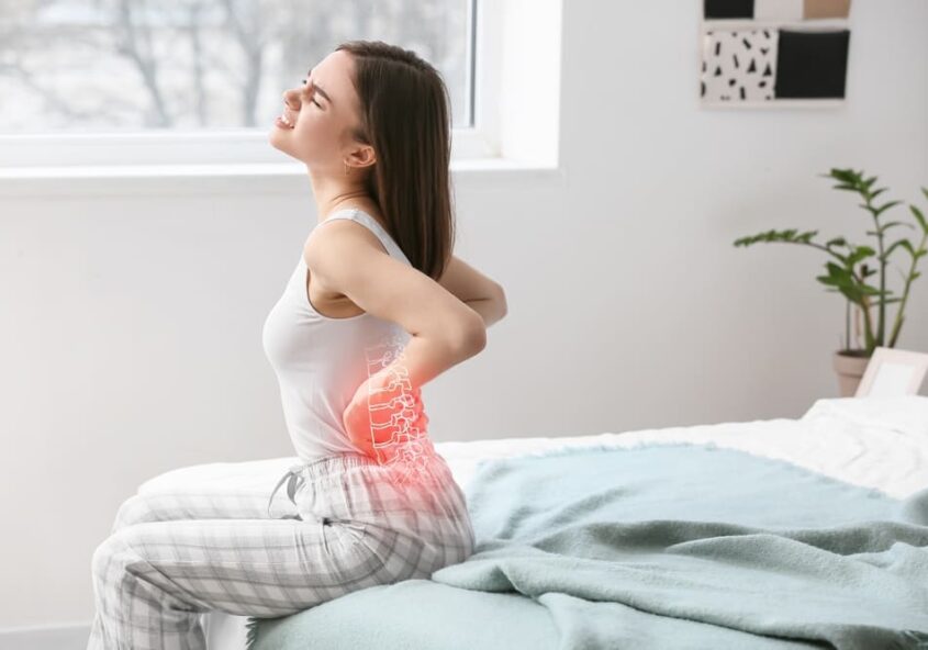 Mujer joven sentada en la cama sujetándose la zona lumbar con dolor, con la columna vertebral resaltada, lo que ilustra el malestar de espalda inducido por el estrés tratado con quiropráctica.