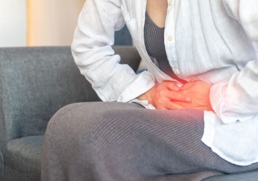Mujer sentada en un sofá agarrándose el bajo vientre con dolor, destacando los síntomas del dolor pélvico crónico tratado en Metro Healthcare Partners Brooklyn.