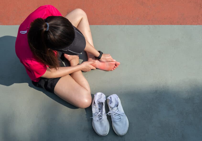 Corredora sentada en la pista sosteniendo y examinando su pie dolorido, ilustrando lesiones comunes en los pies y tobillos de los atletas, como esguinces, fascitis plantar o tendinitis aquilea.