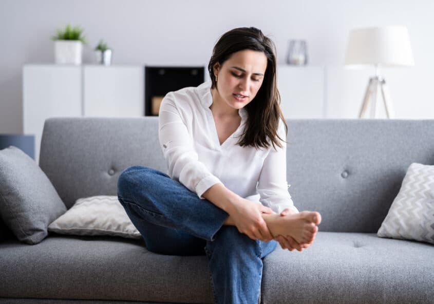 Una joven está sentada en su sofá por la mañana, haciendo muecas de dolor y sujetándose el talón lesionado, un síntoma habitual de la fascitis plantar.