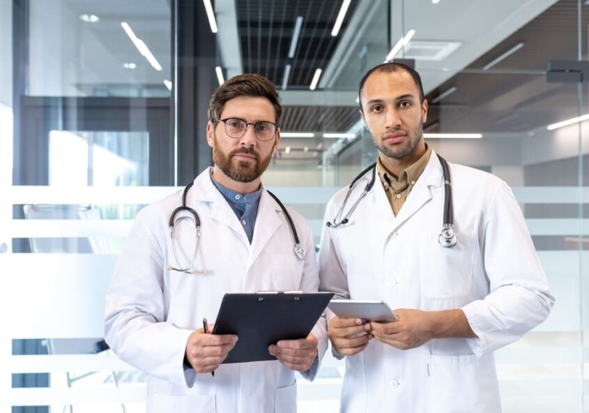 Dos médicos con aire seguro, vestidos con batas blancas y con estetoscopios, de pie en el pasillo de un hospital moderno; uno sostiene una carpeta con sujetapapeles y el otro una tableta, en representación del equipo médico.